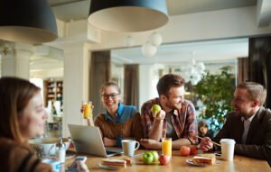 Un groupe de collègues partageant un moment convivial autour d'une table avec des fruits, du café et des ordinateurs dans un bureau lumineux.