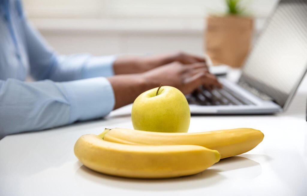 Une pomme verte et deux bananes posées sur un bureau, avec une personne tapant sur un ordinateur en arrière-plan.