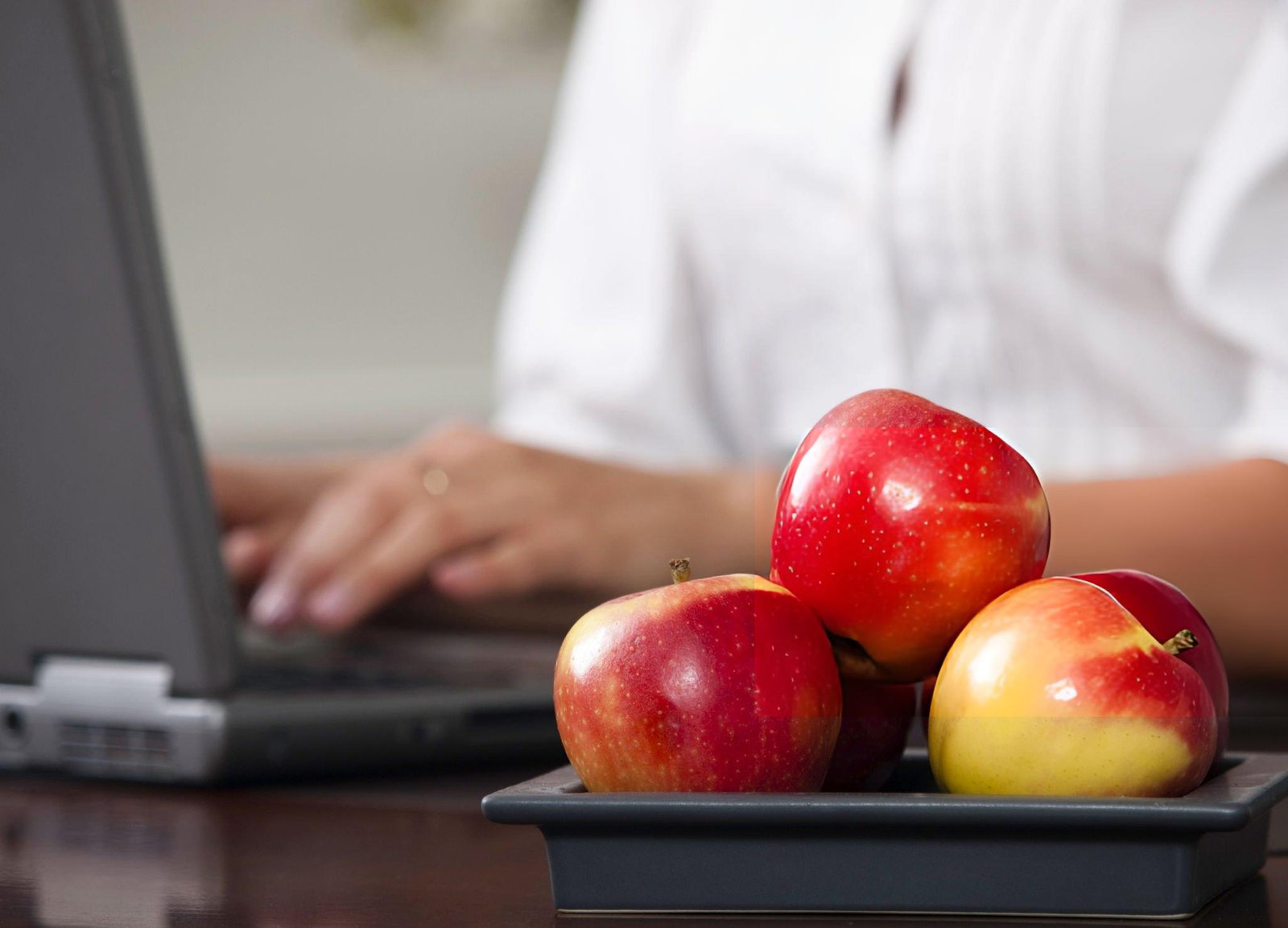 Un bol rempli de pommes rouges sur un bureau, avec une personne travaillant sur un ordinateur en arrière-plan.