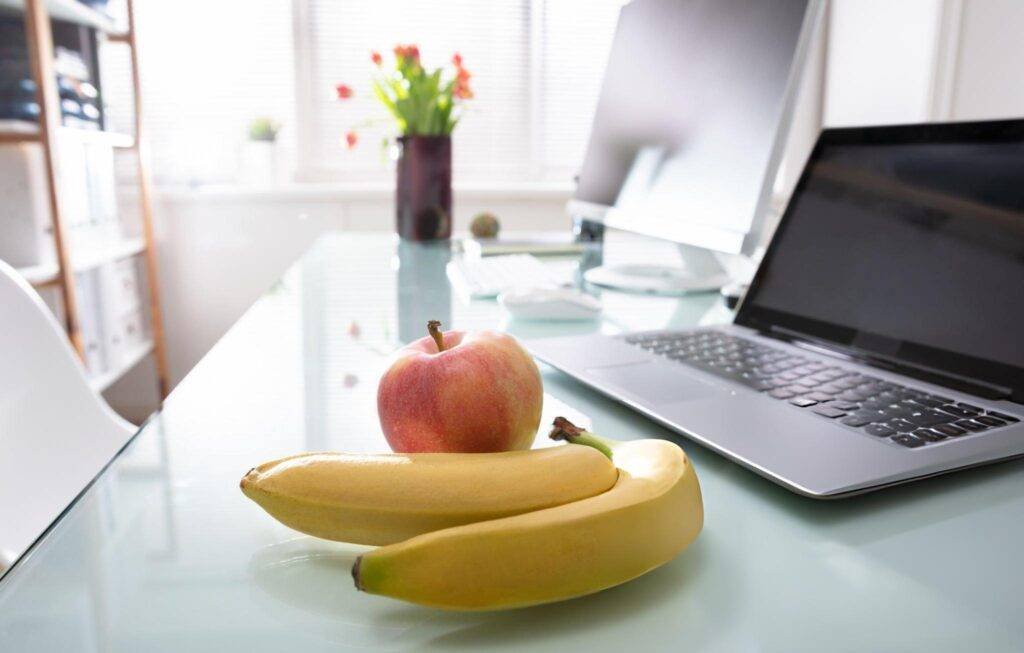 Un bureau minimaliste avec un ordinateur portable sur un bureau en verre, accompagné d'une pomme fraîche et de deux bananes.